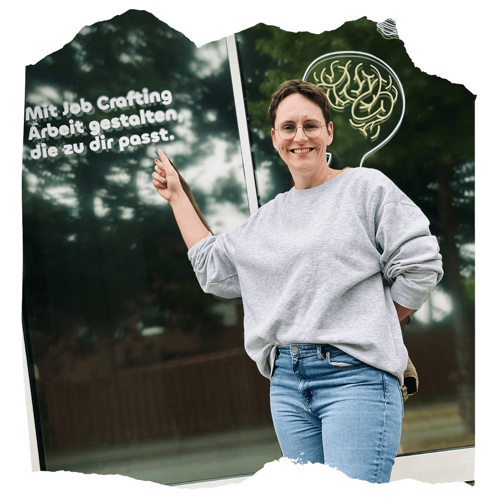 Antonia Ludwig steht vor einem Fenster mit der Aufschrift ‚Mit Job Crafting Arbeit gestalten, die zu dir passt.‘, zeigt darauf und lächelt in die Kamera.